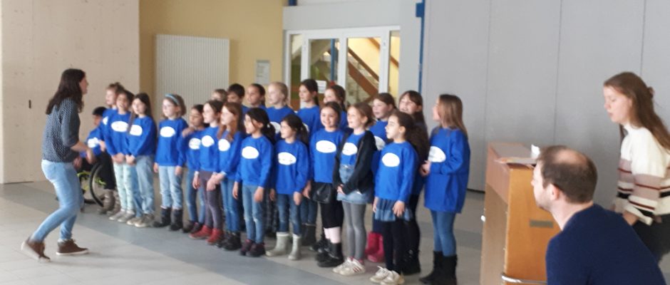 Eine Lehrerin steht vor einem singenden Kinderchor, rechts sitzt eine Frau am Klavier, ein Mann an Cahons