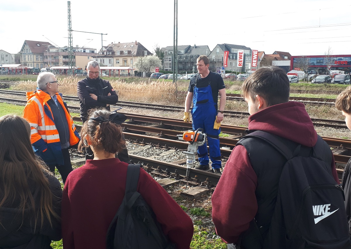 Schüler/-innen hören drei Männern vor Gleisen zu